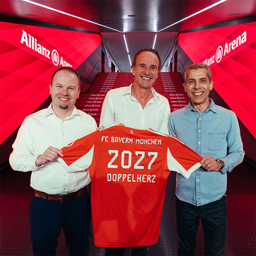 Three men stand in a hallway at Allianz Arena, holding up an FC Bayern München jersey. The jersey is orange with "2027" and "DOPPELHERZ" printed on it. The setting features red lighting, creating a vibrant atmosphere.