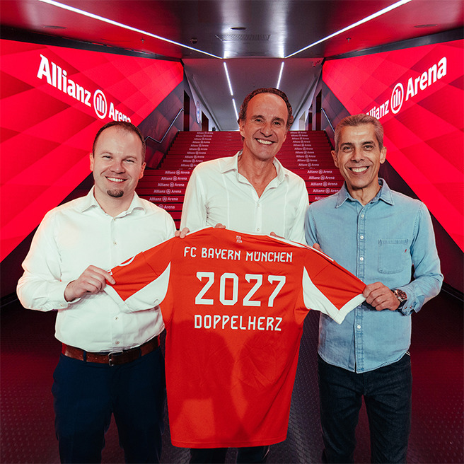 Three men stand in a hallway at Allianz Arena, holding up an FC Bayern München jersey. The jersey is orange with "2027" and "DOPPELHERZ" printed on it. The setting features red lighting, creating a vibrant atmosphere.
