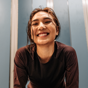 A smiling young woman with wet hair leans forward, appearing joyful and energized. She wears a dark athletic top. The background features light blue panels, contributing to a vibrant atmosphere that evokes a sense of positivity and enthusiasm.