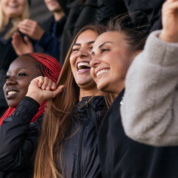 A group of three young women is joyfully interacting, smiling, and celebrating. One woman has long red hair, while another has dark skin and is wearing a black jacket. The third woman has long brown hair and is dressed in a light-colored coat. The atmosphere is lively and cheerful.