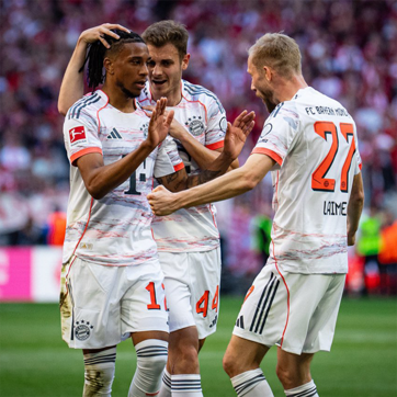 Three Bayern Munich football players celebrate a goal on the field. One player shows joy while another offers support with a hand on his head. Their jerseys feature the team's logo and colors, highlighting a moment of teamwork and excitement during a match.